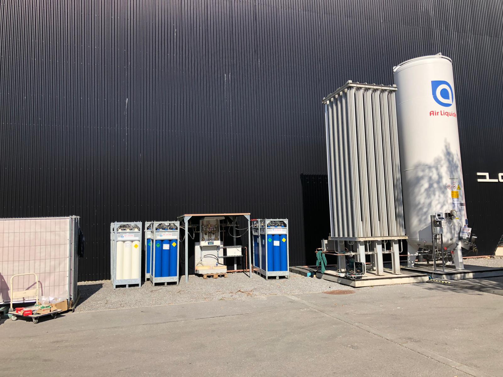 Außenanlage mit Gasversorgungsstation: mehrere Gasflaschenbündel in Gestellen und ein großer vertikaler Lagertank vor der Messehalle Dornbirn.
