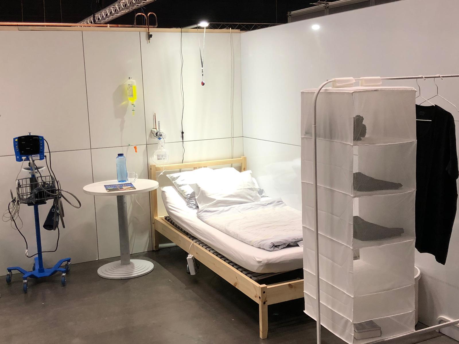 Provisorisches Patientenzimmer in einem modularen Klinikbereich mit Bett, Infusionsständer, Nachttisch und Sichtschutz vor weißen Trennwänden.